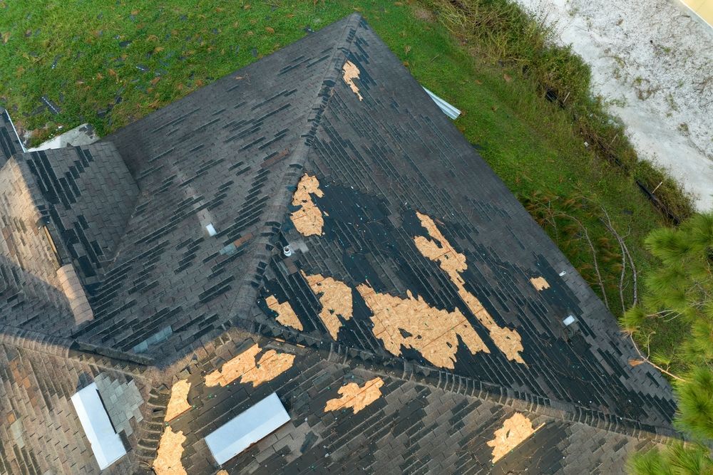 Damaged asphalt shingle roof with missing patches, viewed from above, with green grass in the background.