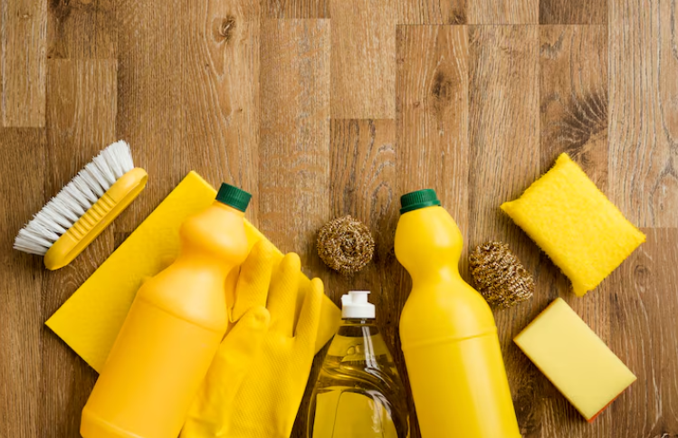 Materiales de limpieza: botellas amarillas, guantes, cepillo para fregar y esponjas sobre suelo de madera.