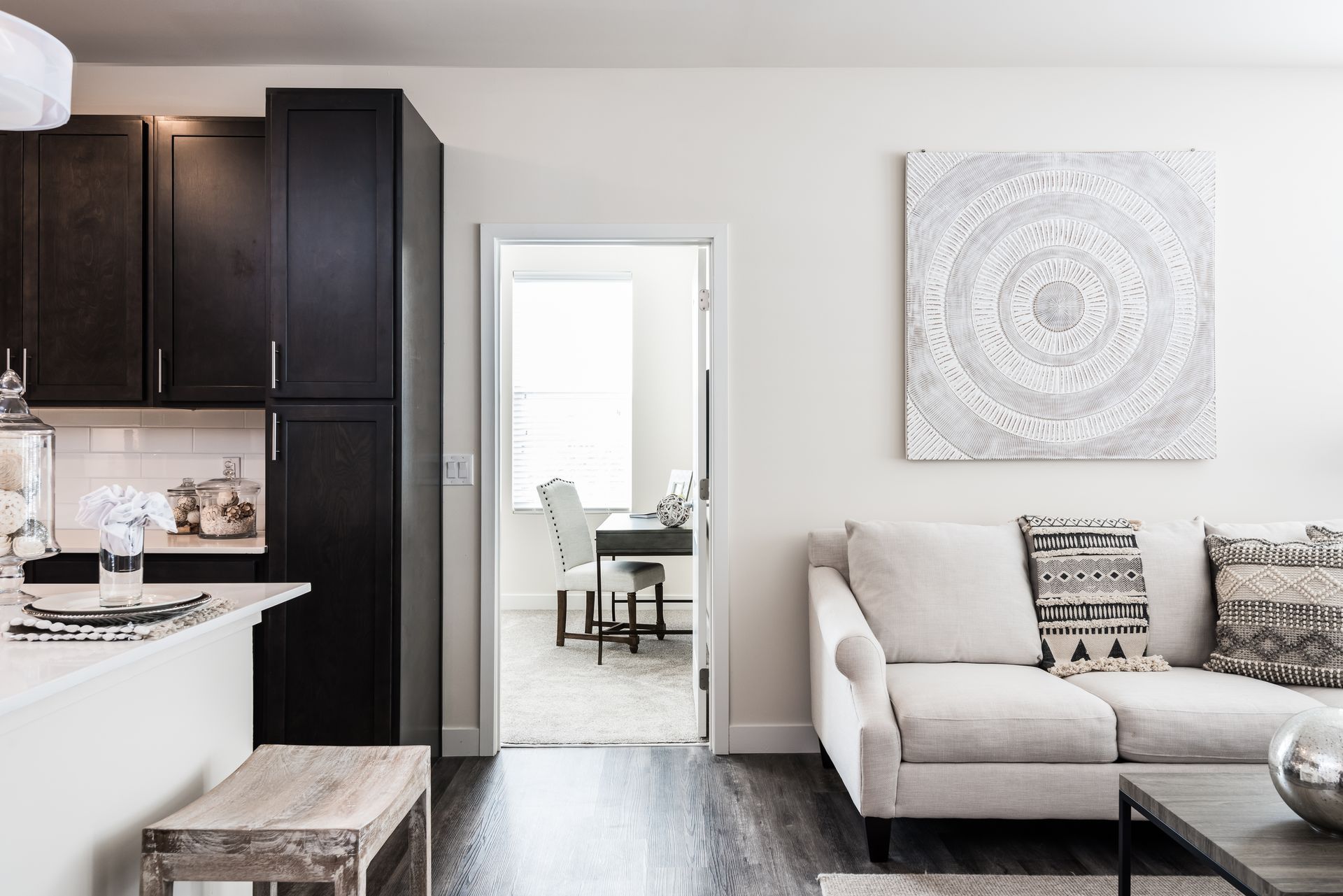 Modern apartment interior with kitchen, living area, and dining room visible. Light walls, dark cabinets, neutral-toned furniture.