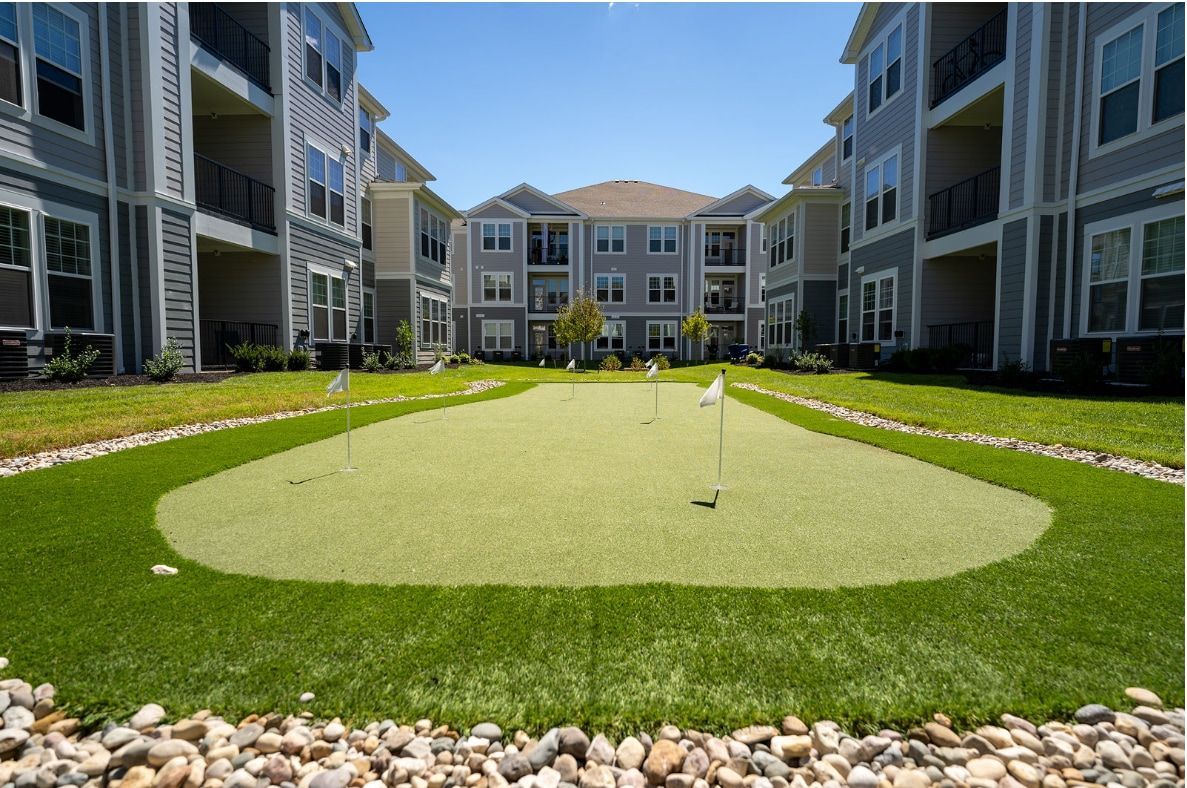 Apartment complex with hammocks strung between trees on a grassy lawn under a clear, blue sky.