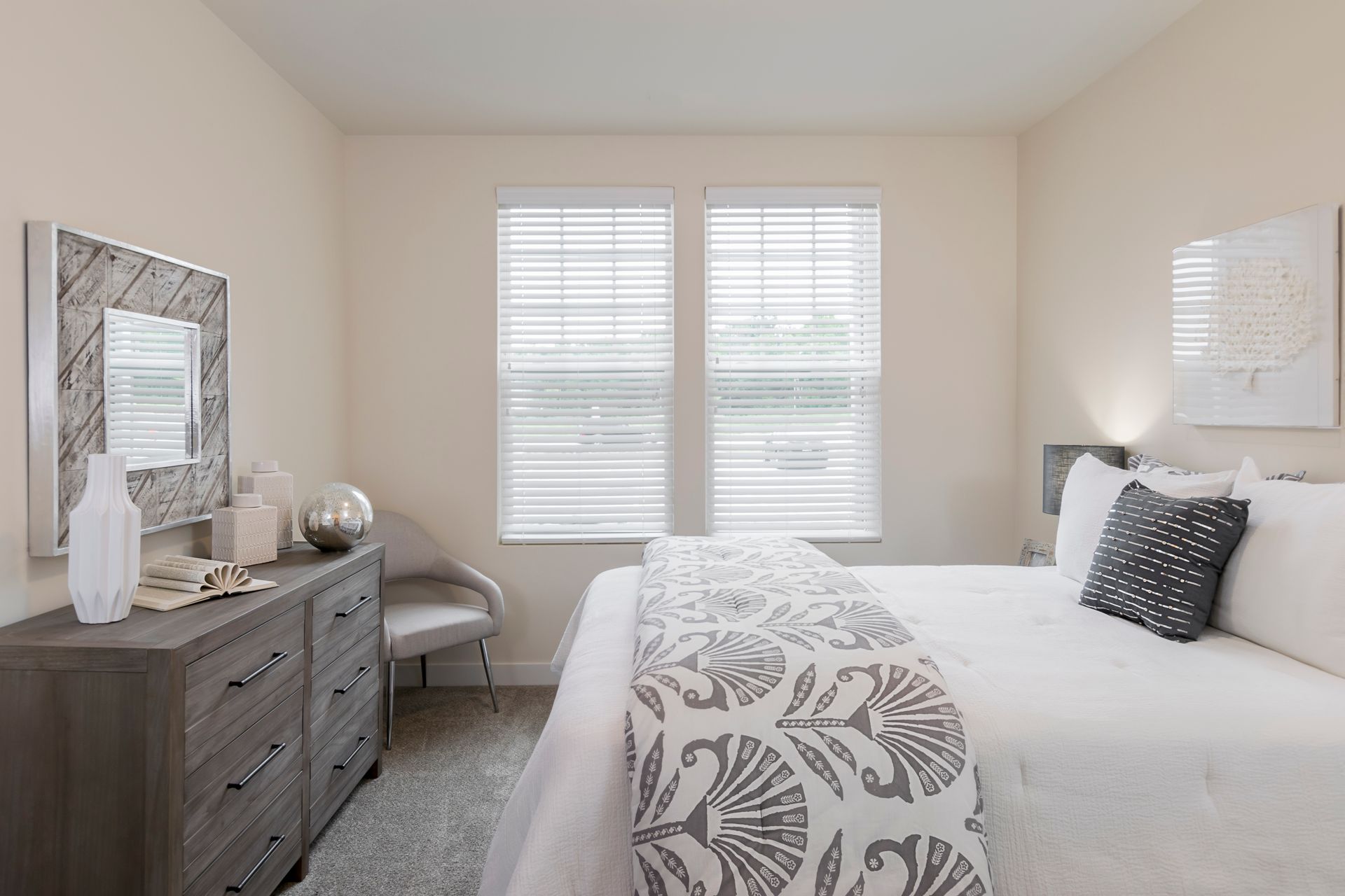 Bedroom with a dresser, bed with decorative pillows, and two windows with blinds.