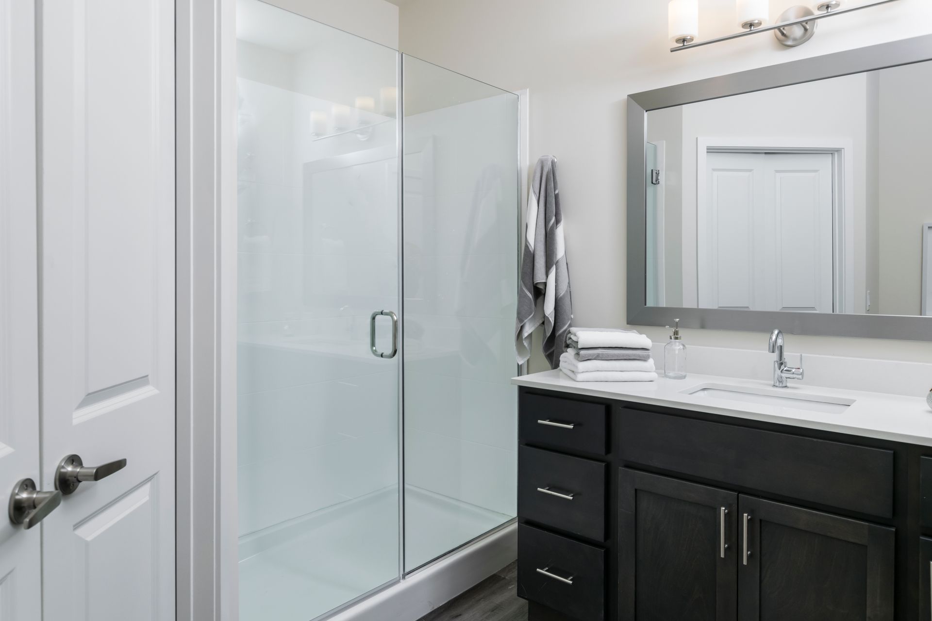 Bathroom with a glass shower, dark vanity, white sink, large mirror, and closed white door.