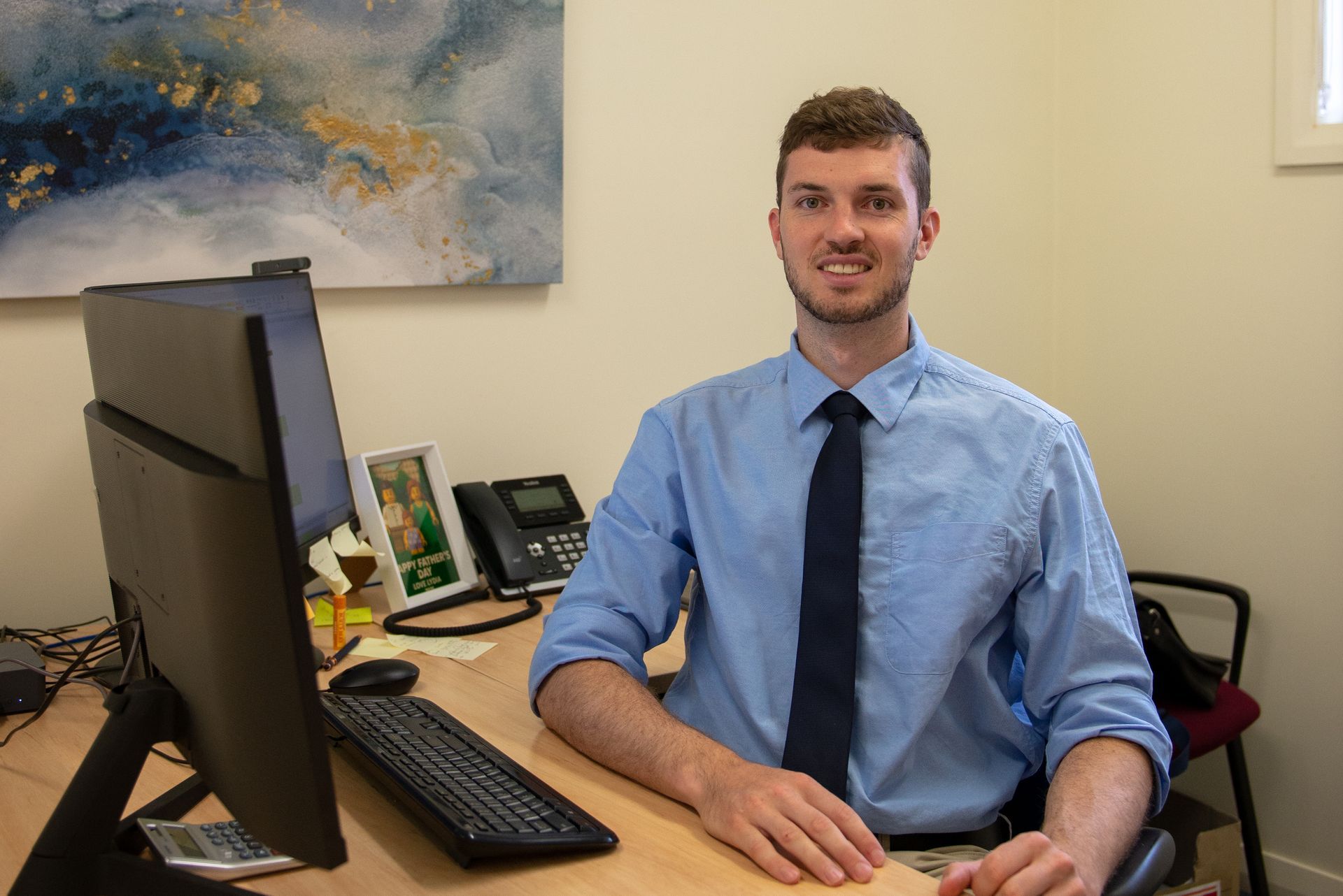 Man at desk in a blue shirt and tie smiles, office setting with monitor, phone, and art — Wright Partners in Dubbo, NSW