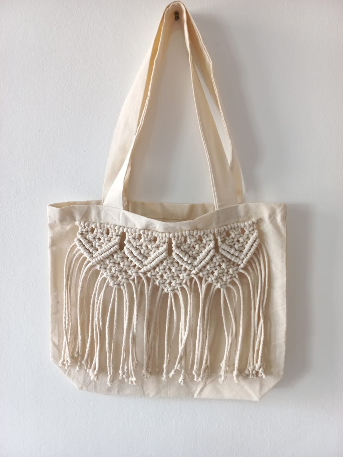 Bolso tote de color crema con detalle de macramé en el frente, colgado sobre una pared blanca.