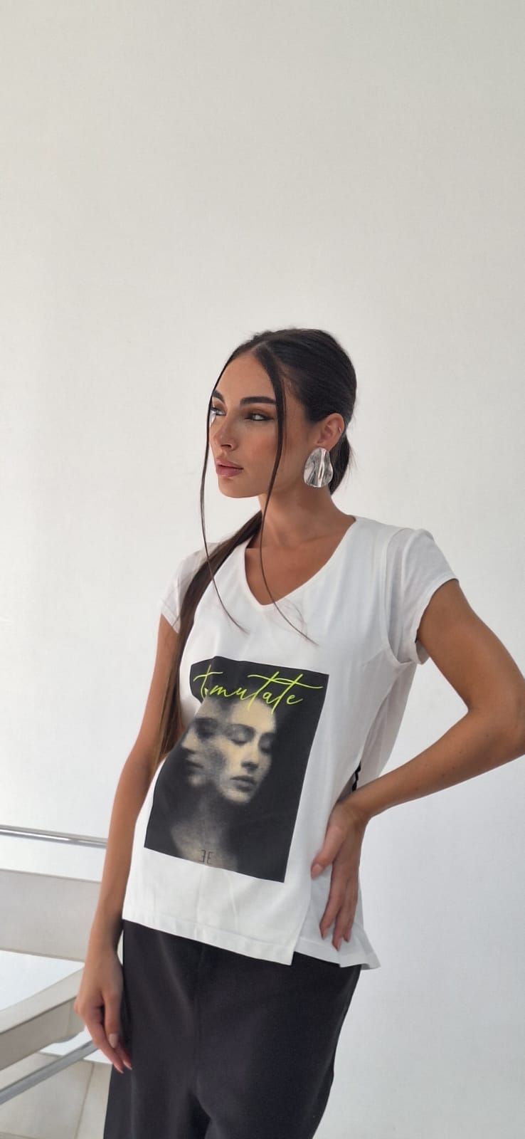 Mujer con camiseta blanca con un dibujo de una cara, sujetando su trenza. Lleva pendientes grandes y pantalón negro.