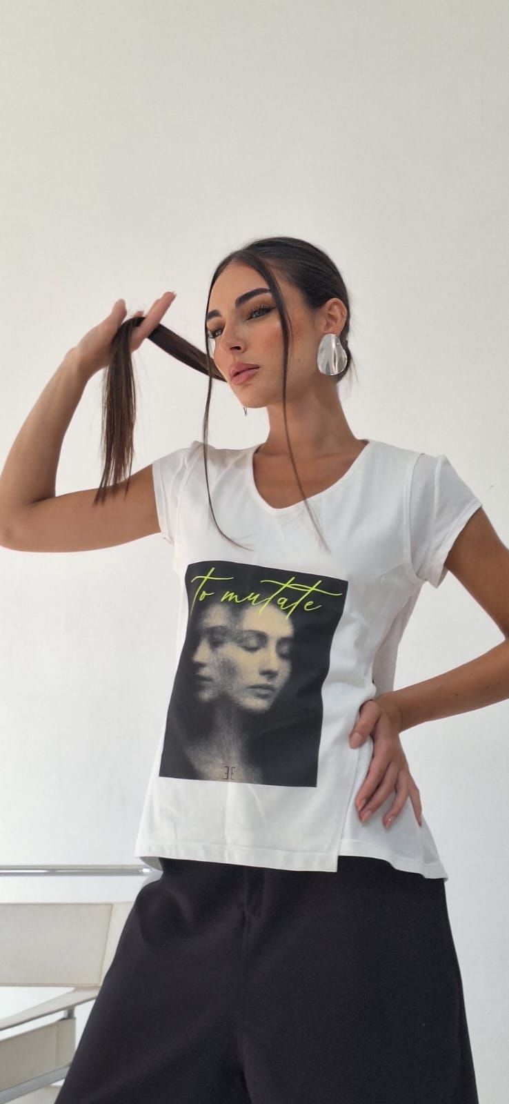 Mujer con camiseta blanca con un dibujo de una cara, sujetando su trenza. Lleva pendientes grandes y pantalón negro.