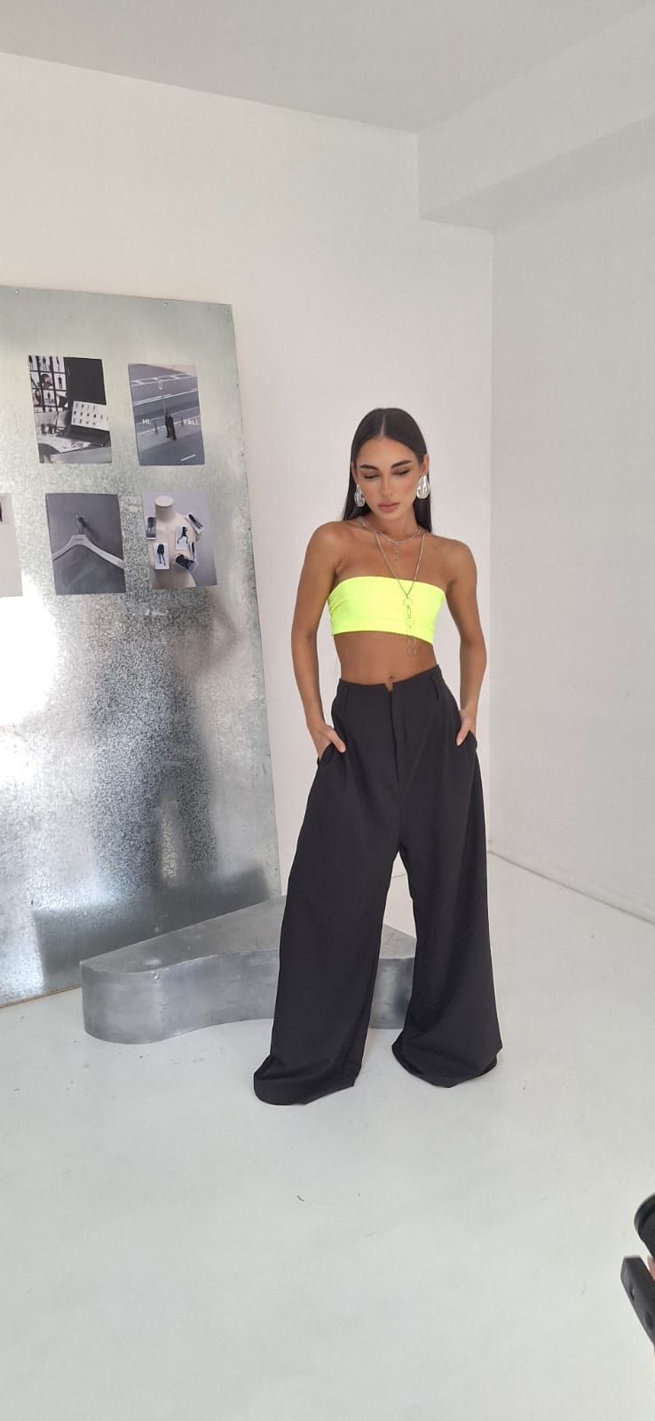 Una mujer con un top de neón y pantalones negros posa con las manos en los bolsillos en un estudio blanco.