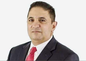 A man in a blue suit, light shirt, and patterned blue tie looking directly at the camera against a white background.