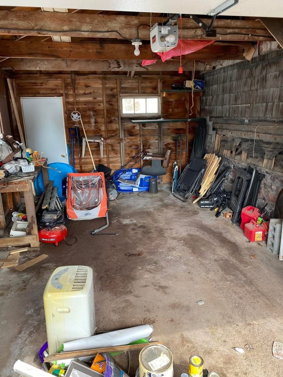 Cluttered garage interior with tools, appliances, and various items. Walls are wood, ceiling beams exposed.