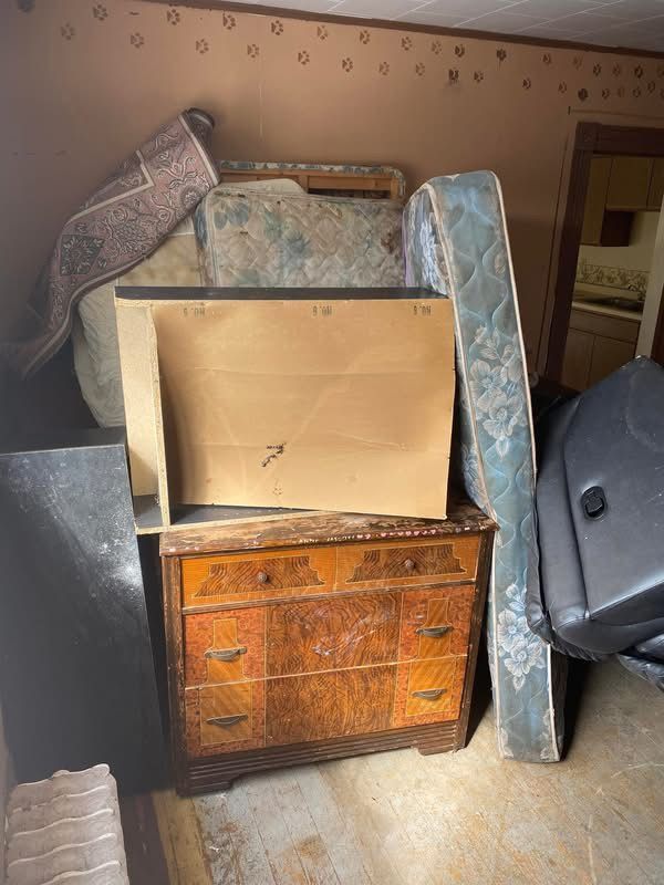 A pile of items in a room, including a dresser, mattresses, rug, and black case, set against patterned wallpaper.