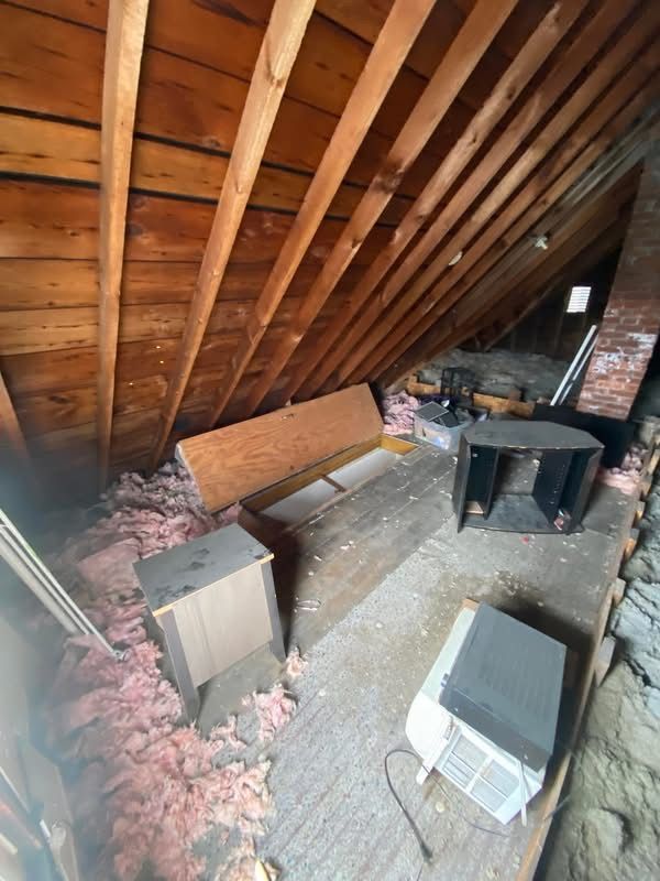 Attic with wood beams, insulation, various objects, brick chimney, and open windows.