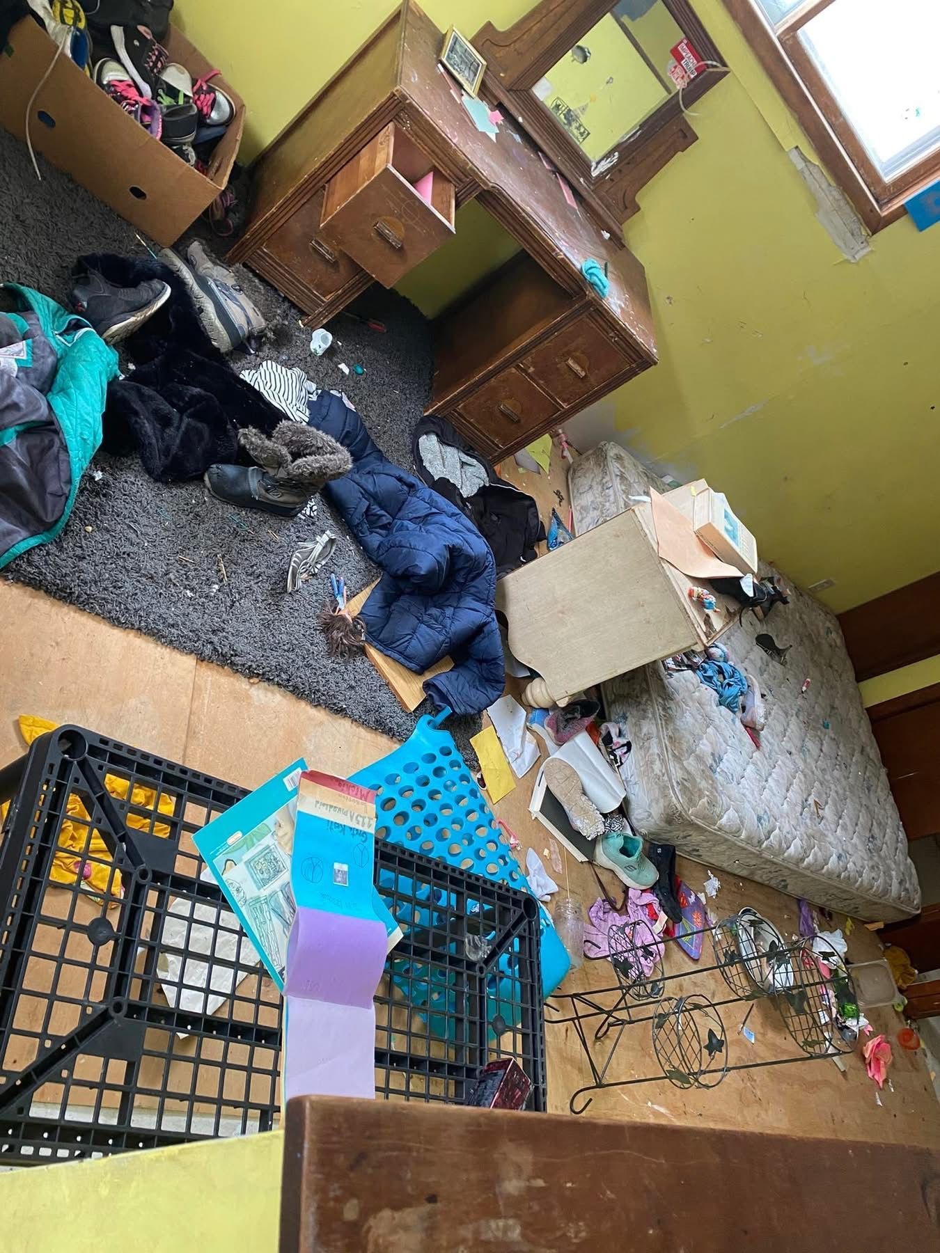 Messy bedroom with clothes, boxes, and furniture scattered on the floor and desk.