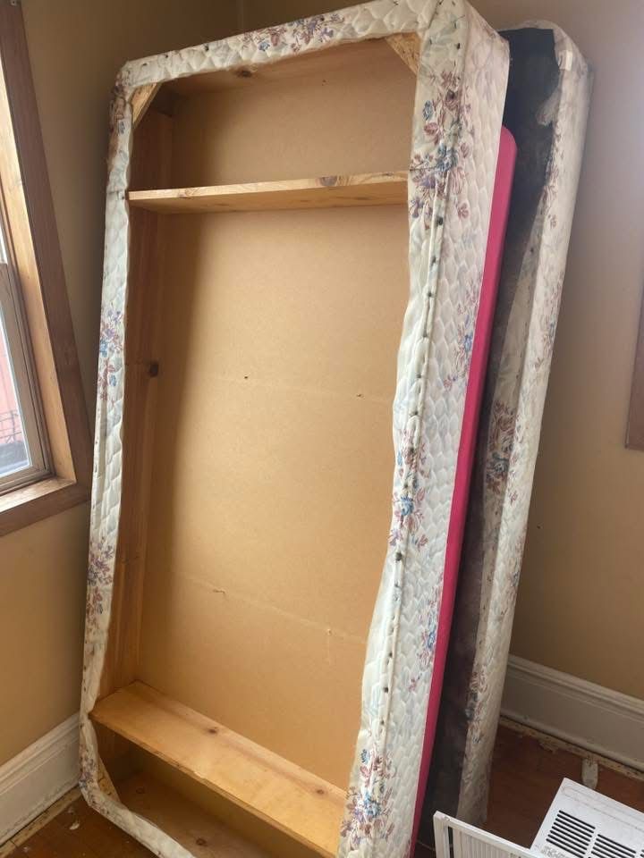 Two mattresses, one with an exposed wooden frame and shelves, leaning against a wall.