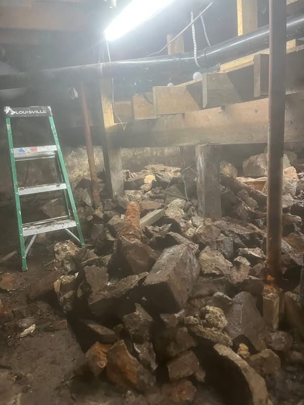 Interior basement with rubble and wooden supports, a green ladder leans against a wall, and pipes run overhead.