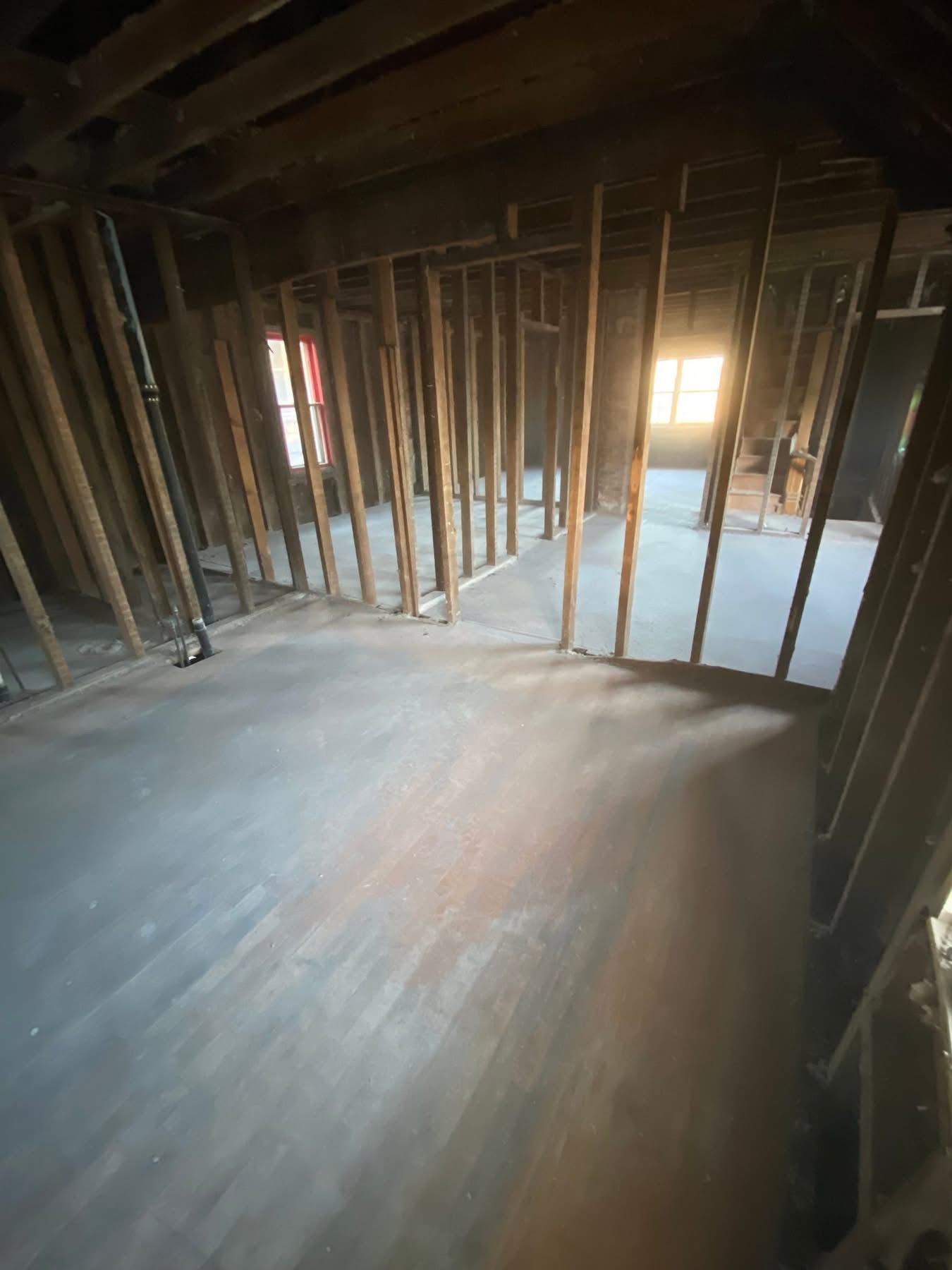 Interior of a room under renovation; exposed wooden framing, dark floor, sunlight through window.