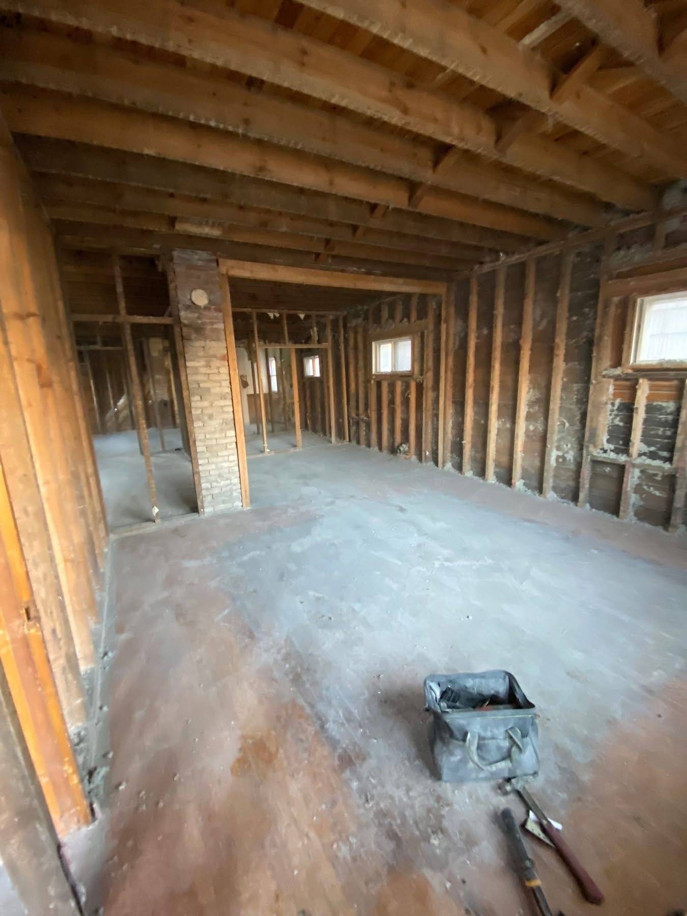 Interior of a room undergoing renovation; exposed wooden beams, studs, and brick chimney; tool bag on floor.
