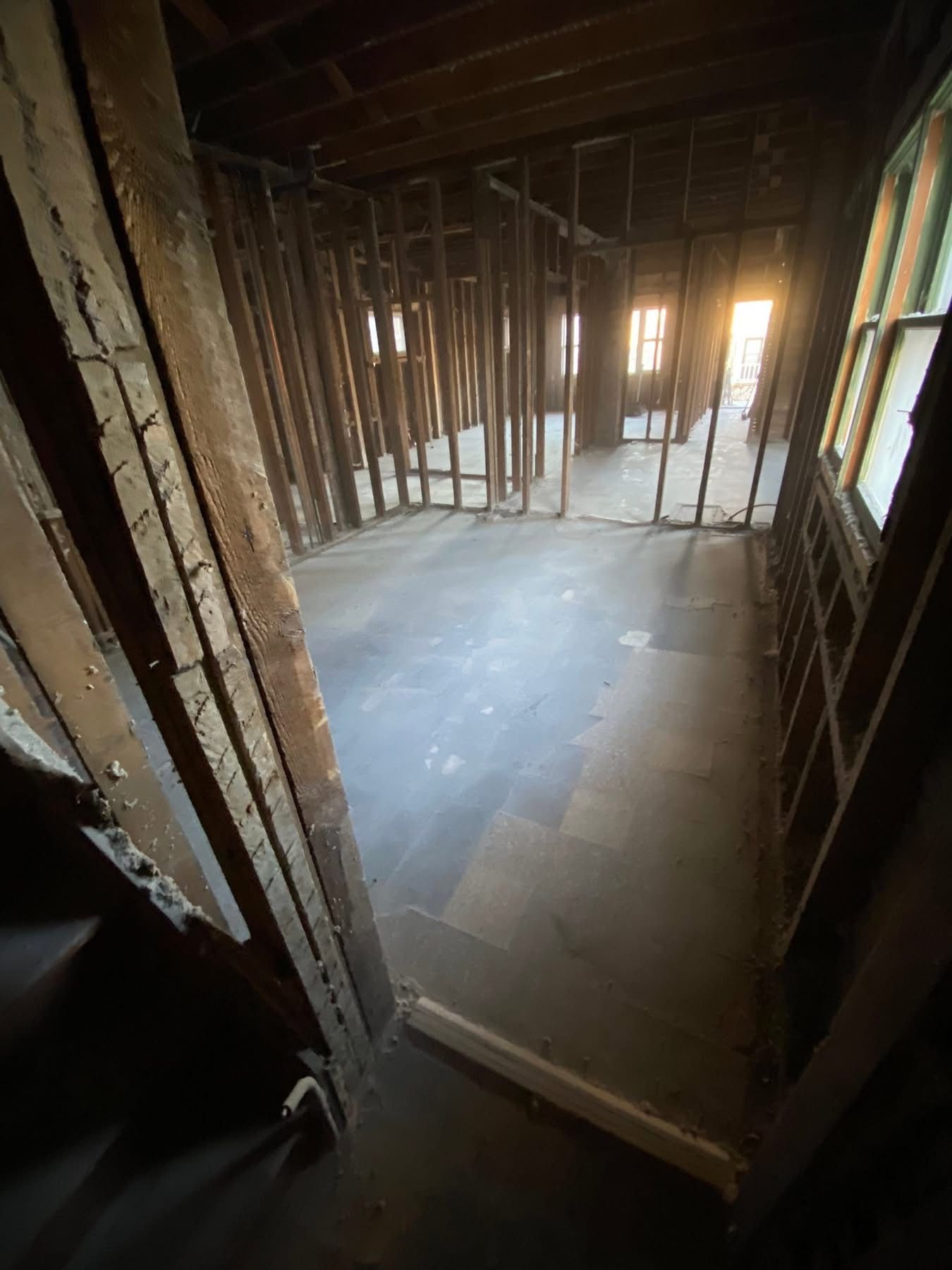 Interior of a room under construction, with exposed wooden studs and partial flooring. Sunlight streams through a window.