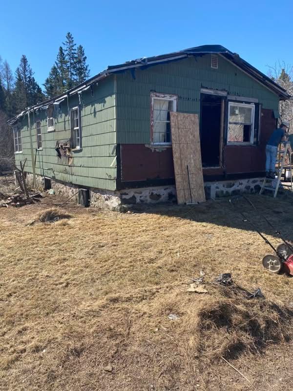 Dilapidated green and maroon house with boarded-up door and windows, in a grassy yard.