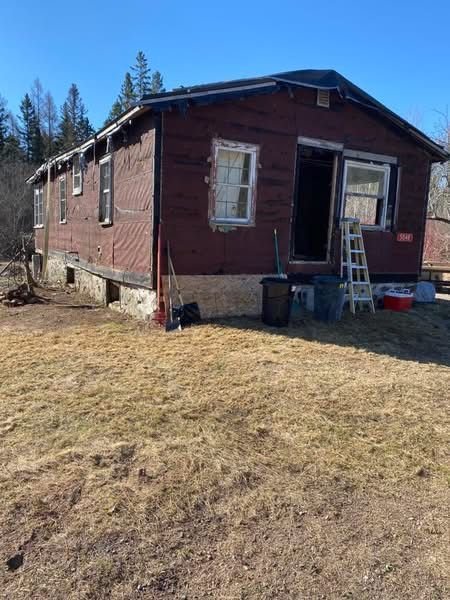 Dilapidated maroon building with open doorway, ladder, and trash cans on dry grass under blue sky.