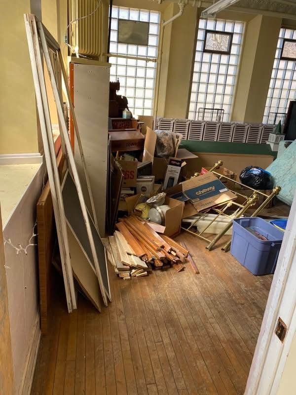 Cluttered room with wooden floors, piles of boxes, furniture, and building materials near windows.