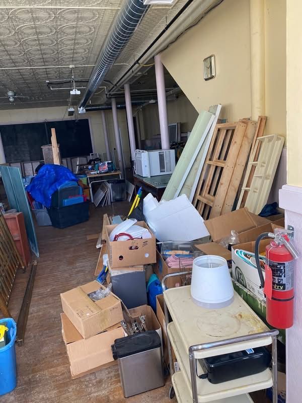 Interior of a cluttered room with boxes, furniture, and a fire extinguisher. Ceiling details and columns.