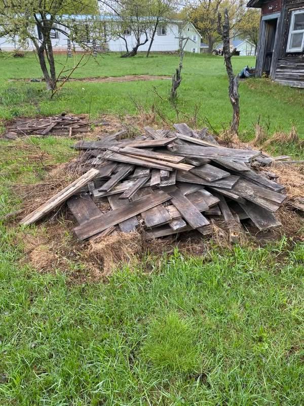 Pile of weathered wooden planks on green grass, next to an old building and a field.