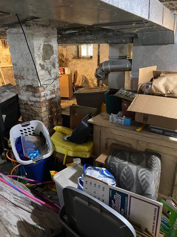 Cluttered basement with boxes, furniture, and a laundry basket under a concrete ceiling and ductwork.