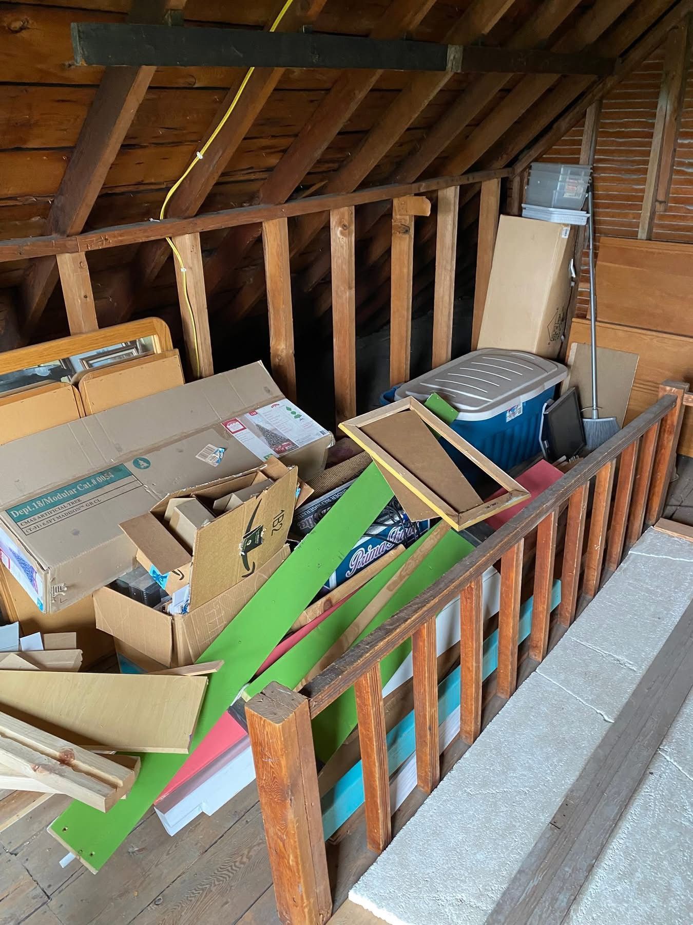 Cluttered attic space with boxes, wood planks, and a blue cooler near a wooden railing.