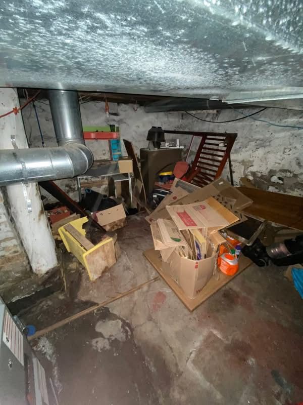 Cluttered basement: boxes, furniture, pipes, concrete floor, ceiling stains.