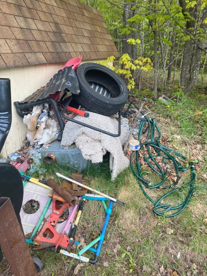 Pile of discarded trash outside a building, including a tire, hose, and chair on grass and leaves.