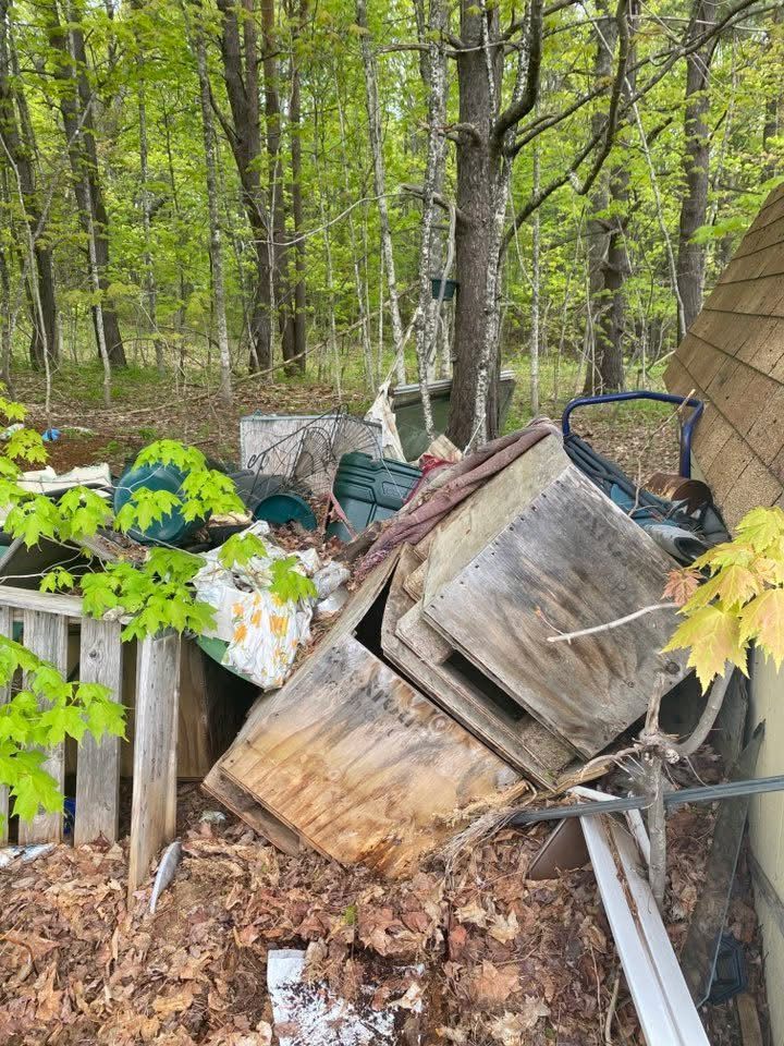 Pile of discarded items: wooden box, cushions, fence, and other debris in a wooded area.
