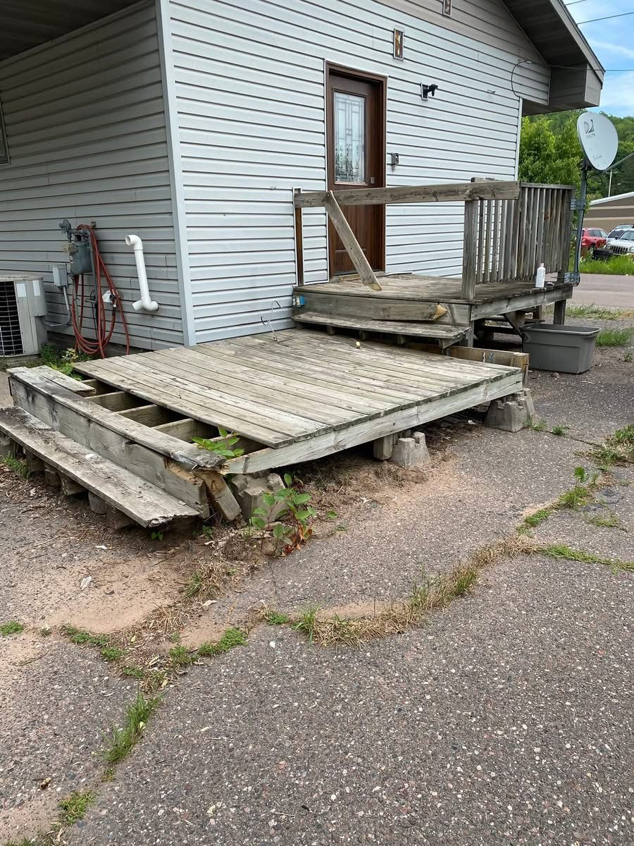 Damaged wooden deck and stairs next to a building with a door, deteriorating with age.