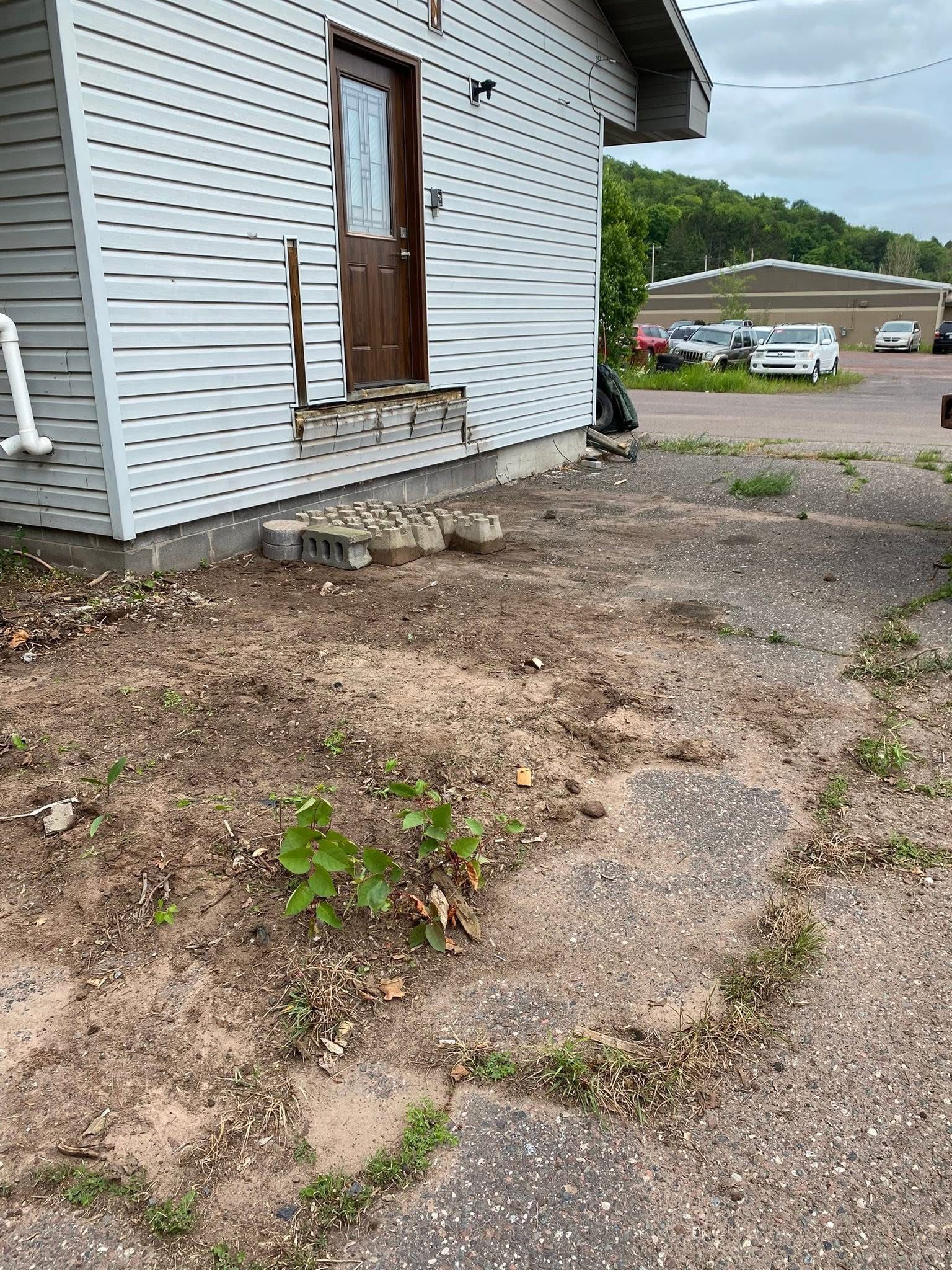 Side view of a building with a door, dirt, a few stepping stones, and sparse vegetation. Cars are visible in the background.