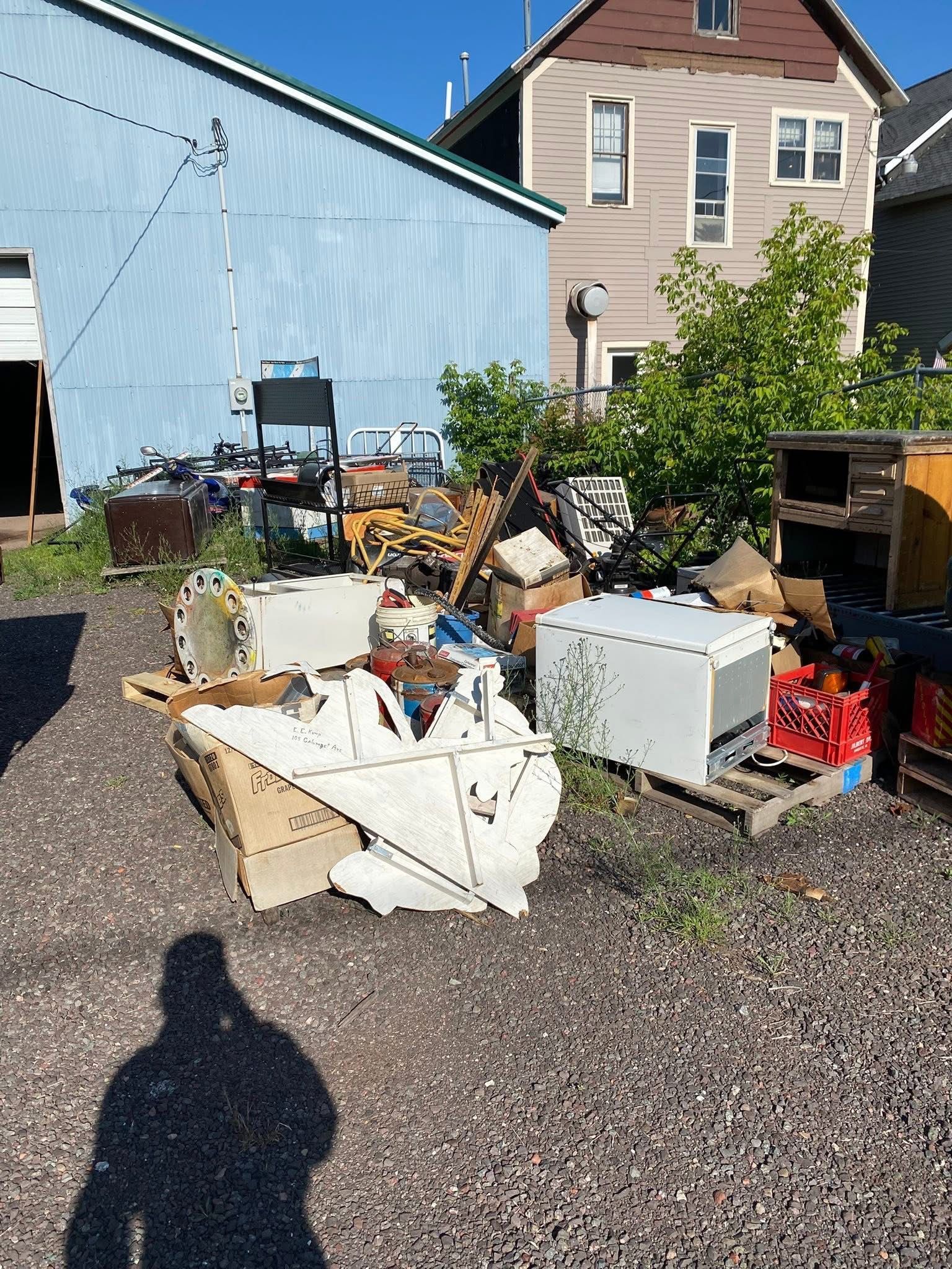 Pile of discarded items, including appliances and wood, in a gravel lot next to a blue building and house.