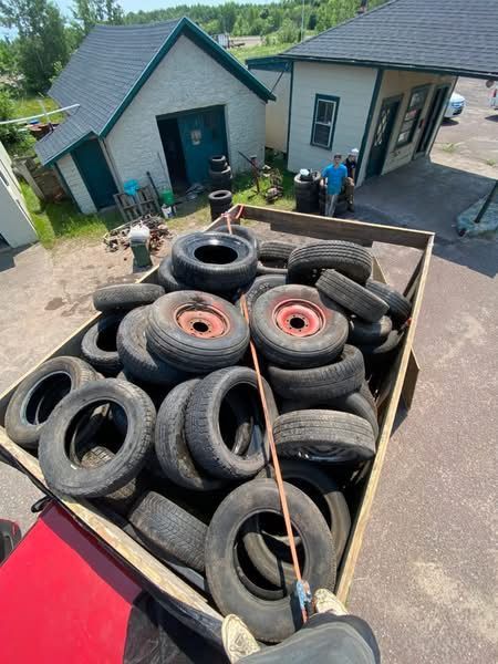 Truck bed overflowing with used tires in front of two small buildings. People in the background.