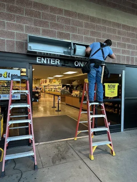 Worker on ladder installing an air curtain above a store entrance with a red ladder nearby.