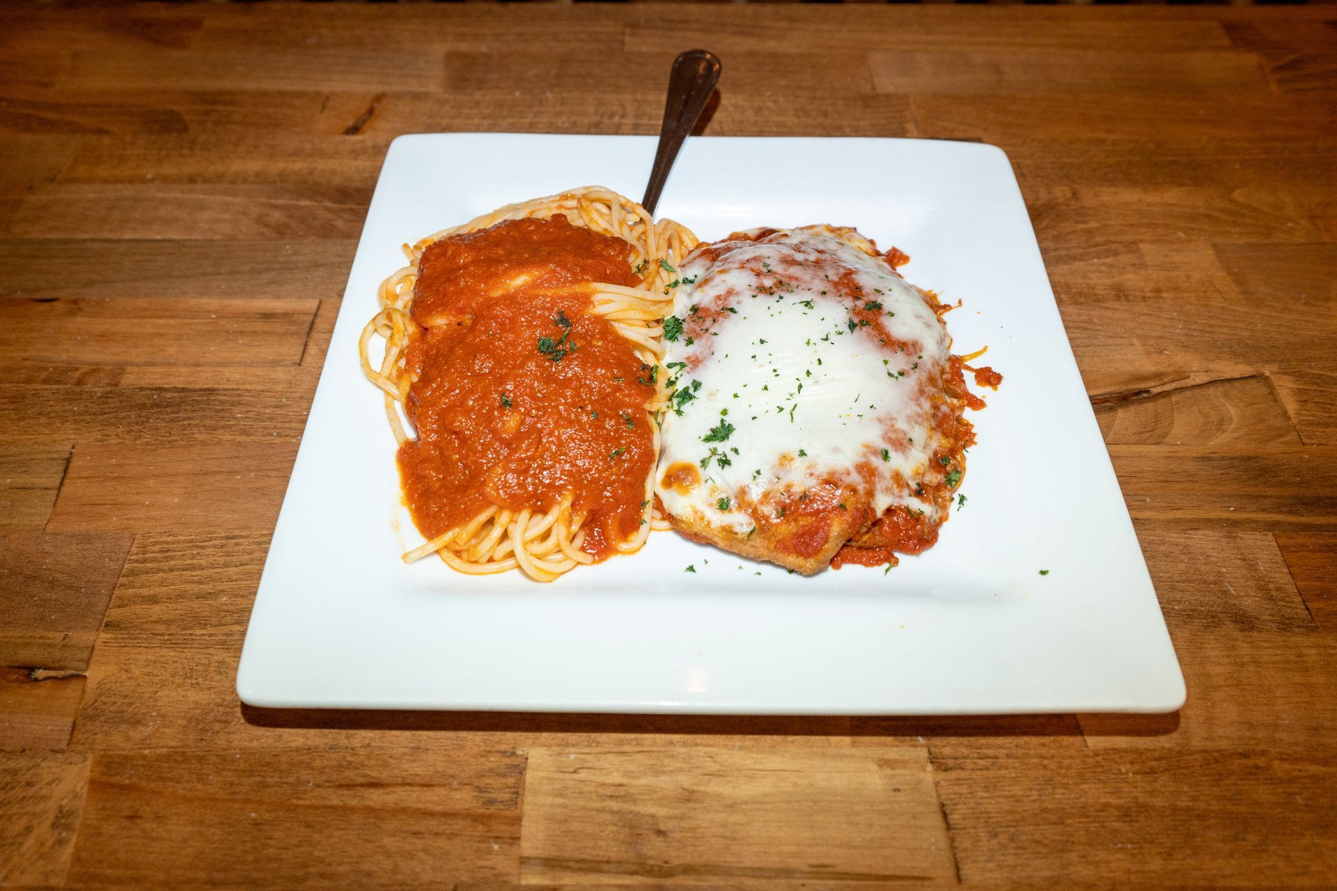 Spaghetti and chicken parmesan on a white plate, with a fork, set on a wooden table.