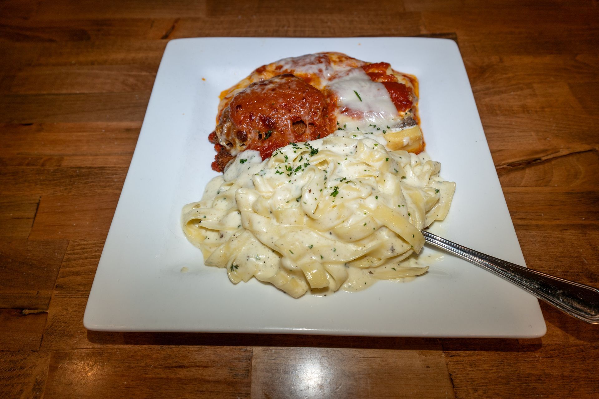 Lasagna with red sauce and cheese, and fettuccine alfredo on a white square plate.