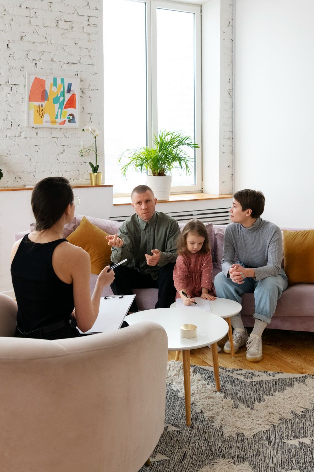 A therapist consults with a family: parents and a child. They are sitting on a couch and the therapist is in a chair, indoors.