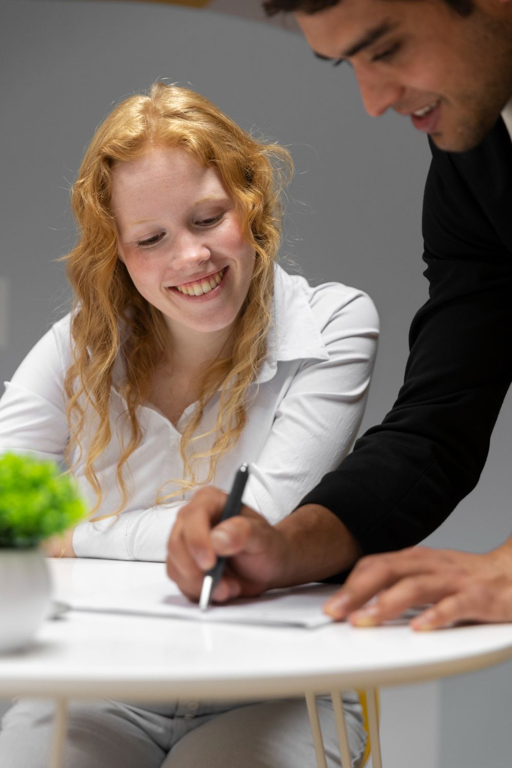 A red-haired woman smiles while a man in a suit leans over her, helping her sign a document at a table.