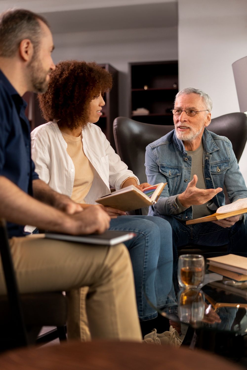 Three people in a meeting: A woman and a man are listening to an older man speaking while holding books. They are in a living room setting.