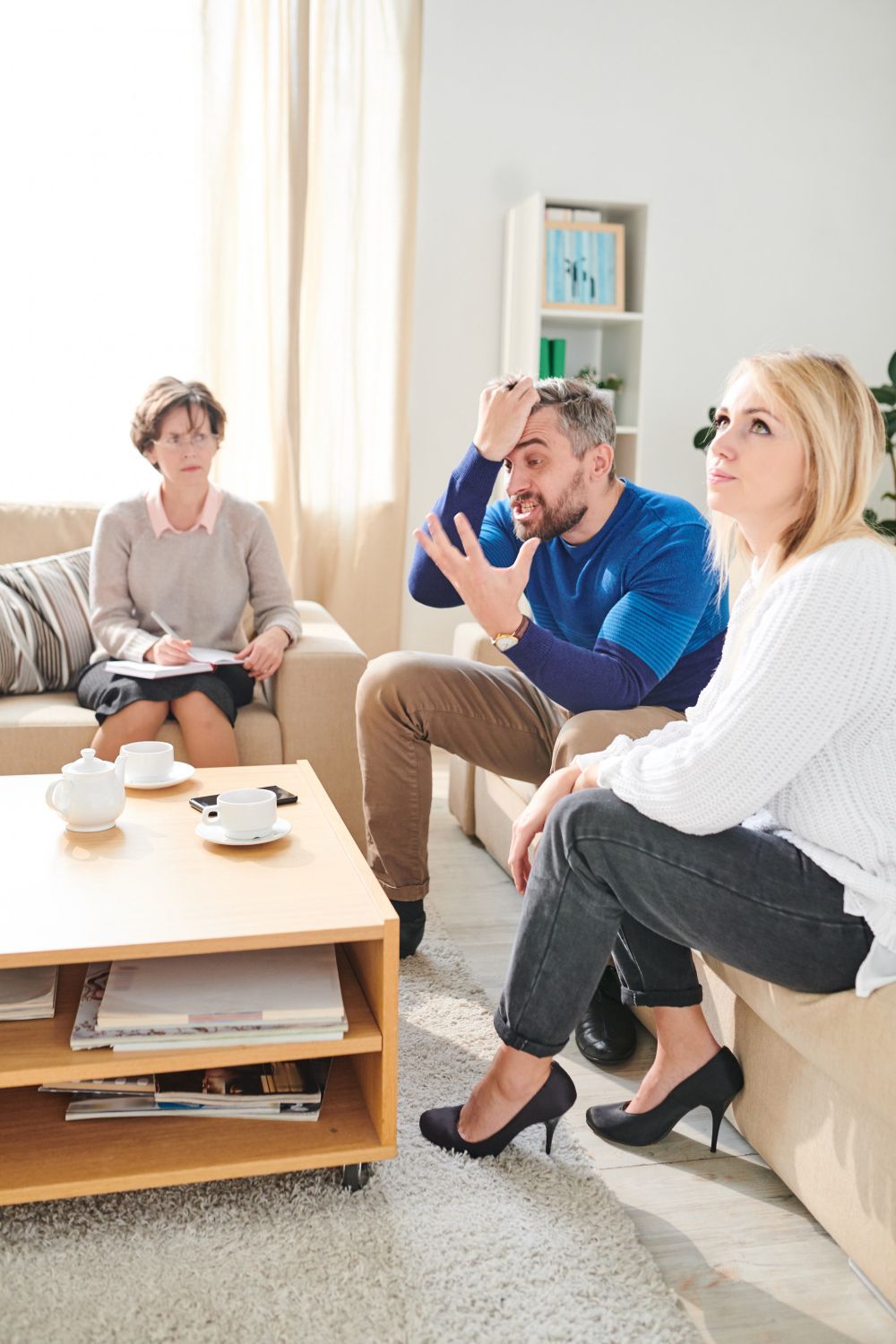 Couple in therapy, man gesturing with frustration, woman looking upward, therapist taking notes on a couch in a brightly lit room.