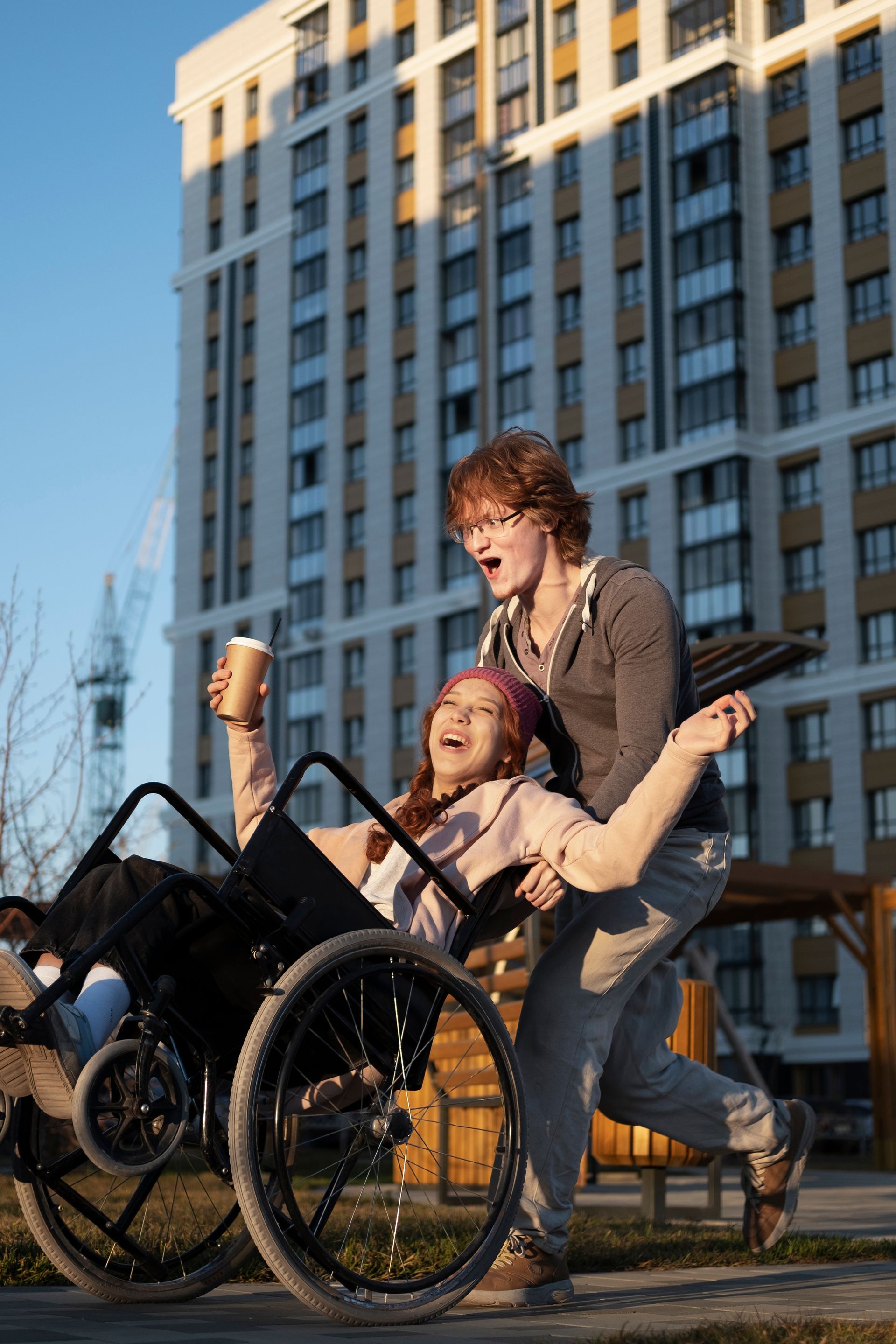 Woman in wheelchair, holding coffee, is being pushed joyfully by a person on a sunny day in front of a tall building.