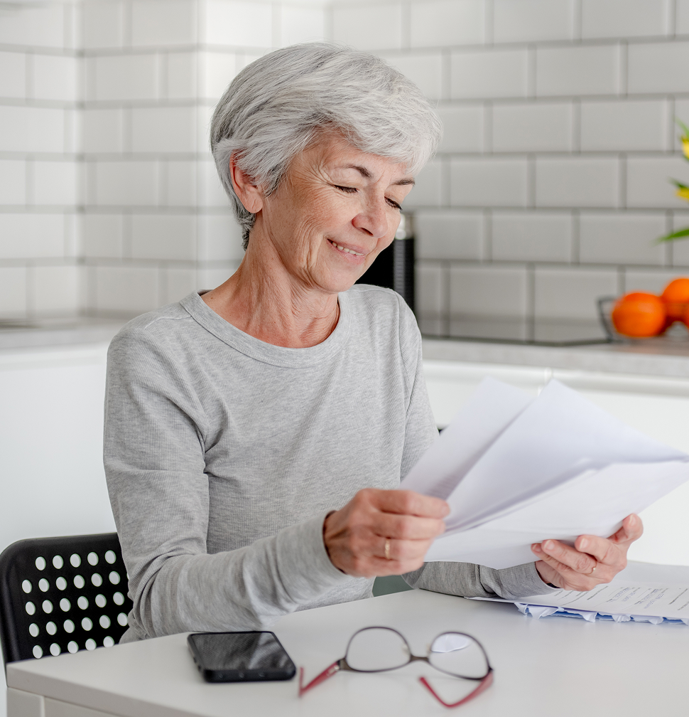 An older woman with gray hair smiles while reviewing documents at a table in a kitchen. A phone and glasses are nearby.
