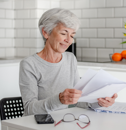 An older woman with gray hair smiles while reviewing documents at a table in a kitchen. A phone and glasses are nearby.