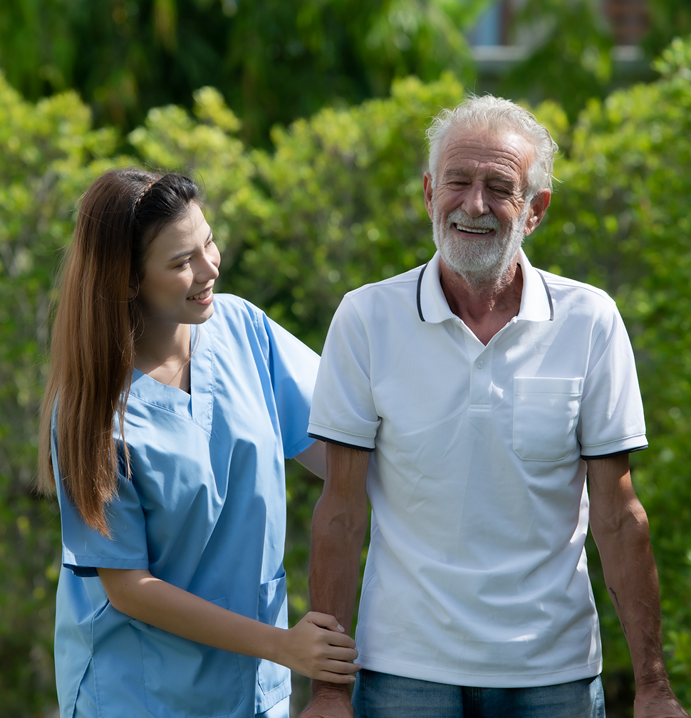 A smiling elderly man walks outside with a young woman in blue scrubs, who is assisting him.