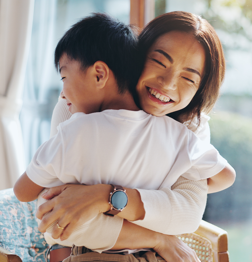 A woman with a wide smile hugs a young boy tightly, both indoors near a window.