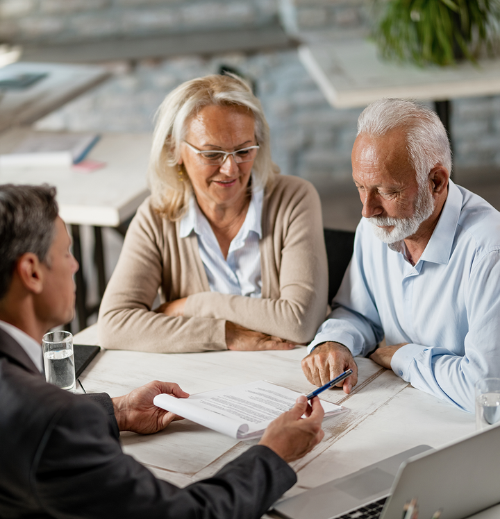 An older couple reviews documents with a financial advisor at a table. They are looking at paperwork, and the advisor is pointing with a pen.