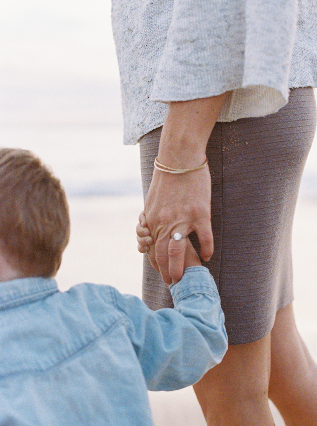 A woman in a skirt and sweater holds a young child's hand on a beach, the child facing away.