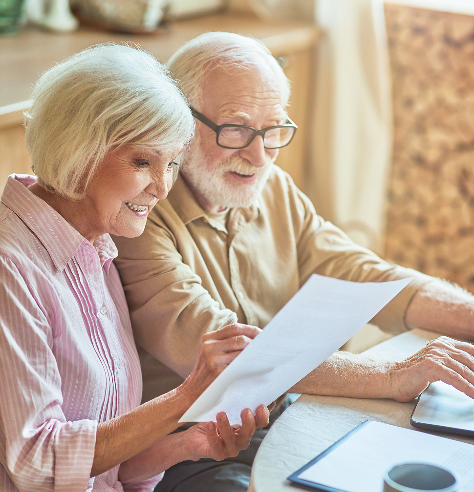 An elderly couple examines paperwork together at a table, one pointing while the other types on a laptop.